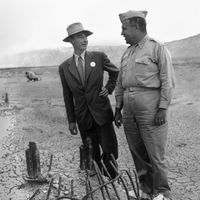 J. Robert Oppenheimer (L) & Gen. Leslie R. Groves at ground zero examine remains of a base of the steel test tower at the Trinity Test site of a nuclear bomb; as part of the Manhattan Project in New Mexico, Sep. 1945. Los Alamos National Laboratory