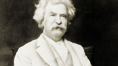 Samuel Clemens aka Mark Twain, three-quarter length portrait, seated, facing slightly right, with cigar in hand.