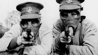 Men of the Royal Norfolk Regiment at Aldershot now undergoing a course of revolver shooting wear gas masks while at practice in order to got used to wearing the masks under all conditions. Two Tommies sighting the target in their gas masks. (World War I)