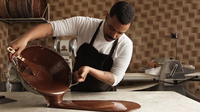 Professional chef pours tasty melted chocolate from one big steel pot to vintage marble table