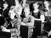 Vintage image of students at desks in a classroom with a teacher standing in the background. (education, learning)